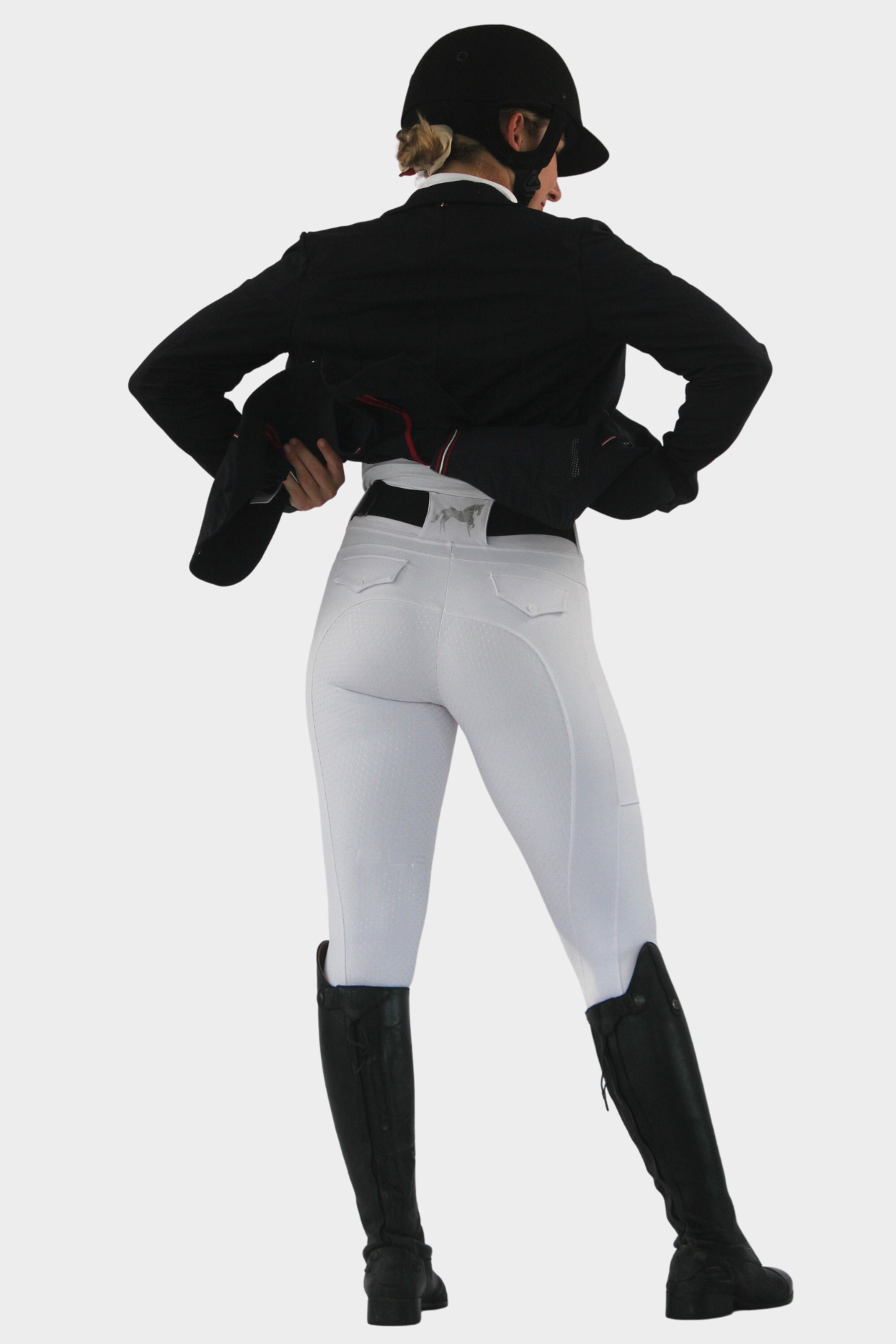 A person wearing a black equestrian helmet, black jacket, white riding breeches, and black riding boots is facing away from the camera, holding the bottom of their jacket with both hands. The background is white. The Pure Canter Competition Tights - White feature a silicone grip for enhanced stability.