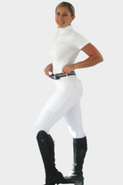 A person stands against a plain background, smiling, and wearing an all-white equestrian outfit crafted from performance fabric. The ensemble includes a short-sleeve shirt, fitted riding tights with silicone grip called Competition Tights - White by Pure Canter, and tall black riding boots. They have their hands on their waist, posing confidently.