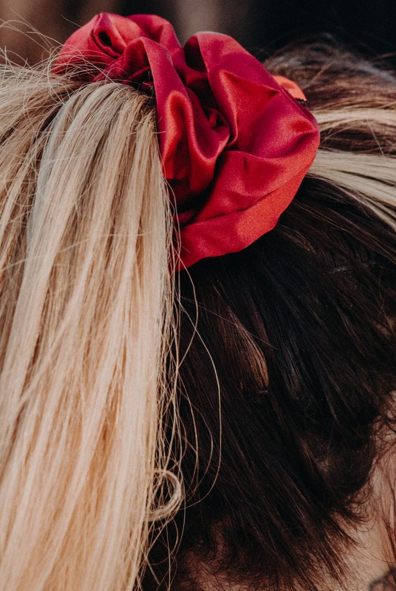 Close-up of a person's hair tied into a ponytail with an Oversized Scrunchie from Pure Canter Pty Ltd. The hair is blonde with darker roots visible. The person's ear is adorned with multiple earrings, perfect for casual outings. The background is blurred.
