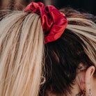 Close-up of a person's hair tied into a ponytail with an Oversized Scrunchie from Pure Canter Pty Ltd. The hair is blonde with darker roots visible. The person's ear is adorned with multiple earrings, perfect for casual outings. The background is blurred.