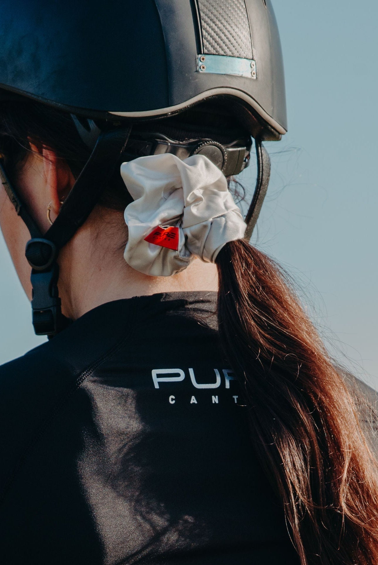 Person wearing a black helmet and a black shirt with "PUR" printed on it. Their long hair is tied back with one of those beautiful **Pure Canter Pty Ltd Oversized Scrunchies**, and they are facing away from the camera against a blurred, outdoor background.
