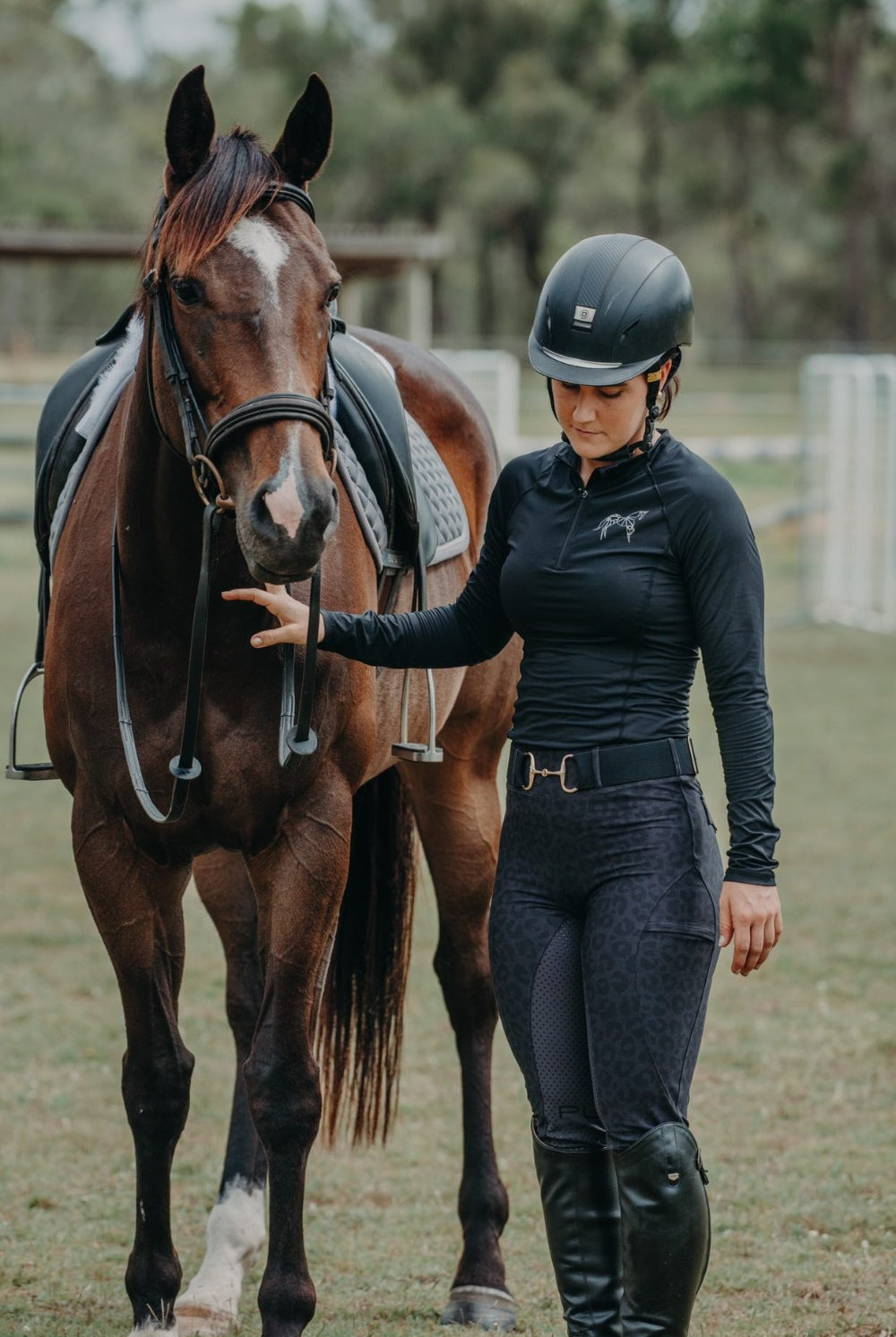 A person dressed in equestrian attire, including a helmet, long-sleeved shirt made of performance fabric, and Fusion Tights - Black Leopard by Pure Canter riding boots, stands beside a brown horse. The person is holding the horse's reins while gently touching its neck. They are outside on a green field with trees and fences in the background.