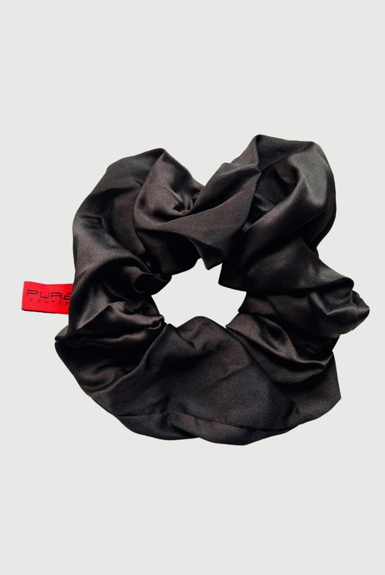 A beautiful Oversized Scrunchie made of shiny black fabric with a subtle gathered texture. Attached to it is a small red label with text that reads "PURE." Perfect for casual outings, the background is plain grey. This exquisite accessory is brought to you by Pure Canter Pty Ltd.