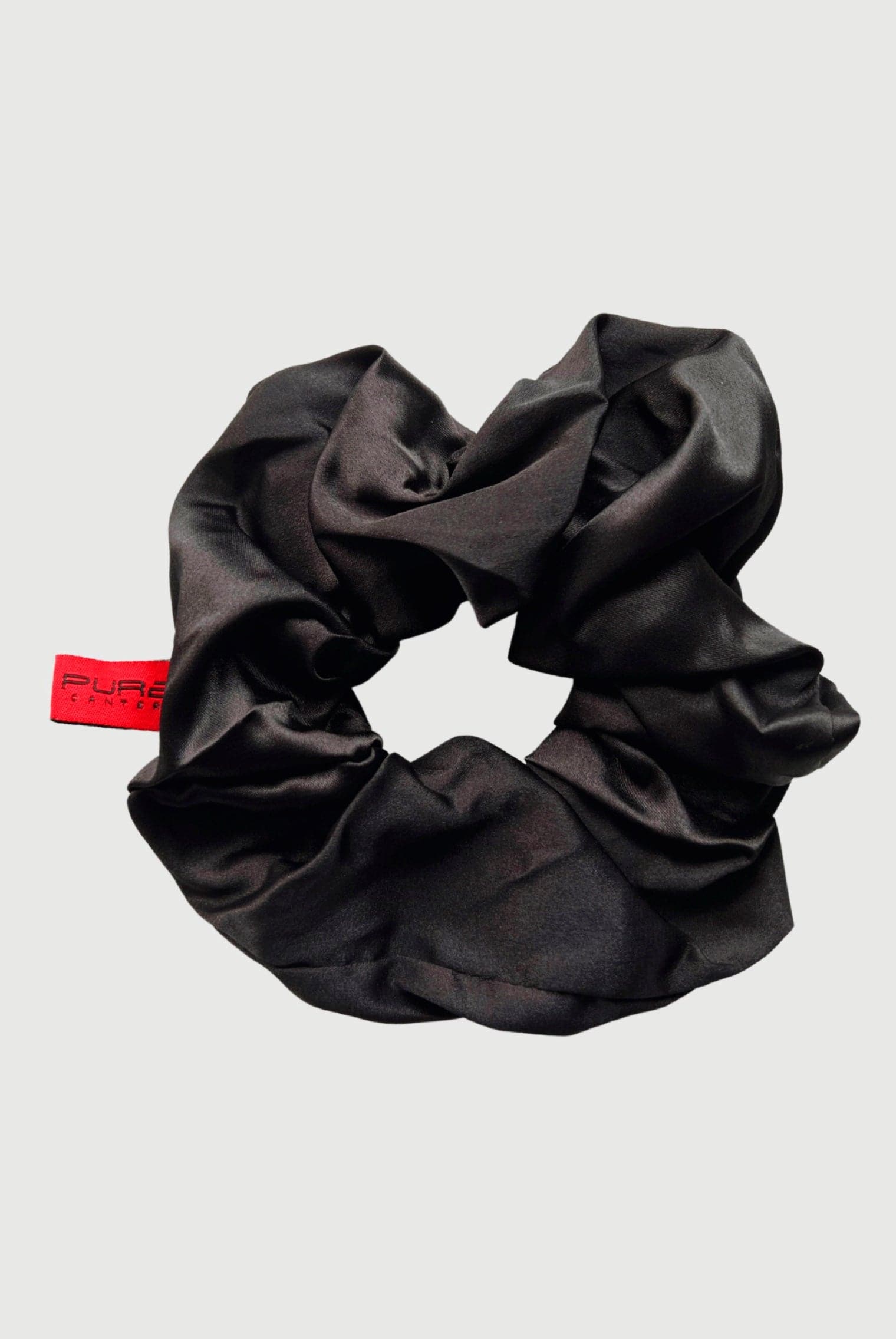 A beautiful Oversized Scrunchie made of shiny black fabric with a subtle gathered texture. Attached to it is a small red label with text that reads "PURE." Perfect for casual outings, the background is plain grey. This exquisite accessory is brought to you by Pure Canter Pty Ltd.