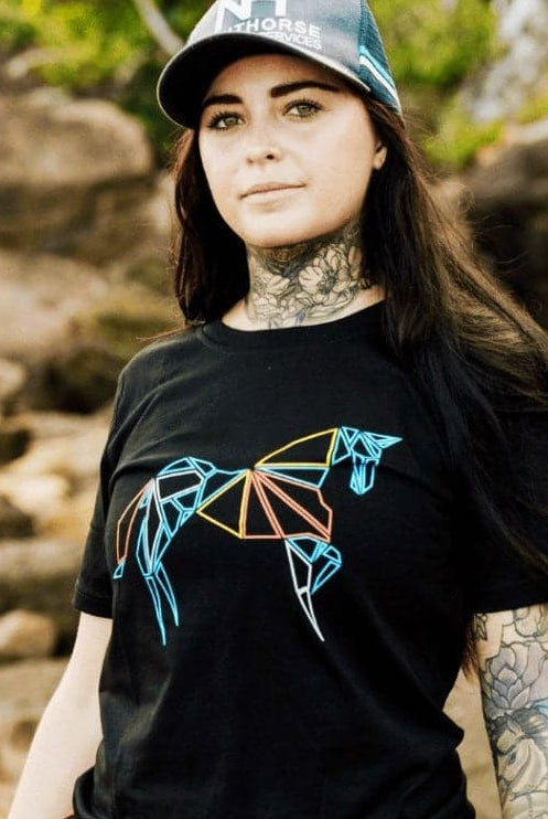 A person with long dark hair, tattoos, and a cap stands on a rocky outdoor terrain. They are wearing a black Youth PURE Emblem Shirt by Pure Canter with a colorful, abstract geometric horse design on the front—a true wardrobe staple.