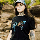 A person with long dark hair, tattoos, and a cap stands on a rocky outdoor terrain. They are wearing a black Youth PURE Emblem Shirt by Pure Canter with a colorful, abstract geometric horse design on the front—a true wardrobe staple.