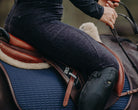 Close-up of a person riding a horse, showing the rider's lower body, saddle, and horse's back. The rider is wearing dark Pure Canter Fusion Tights - Black Leopard and black riding boots. The saddle is brown leather with a dark blue quilted saddle pad underneath.