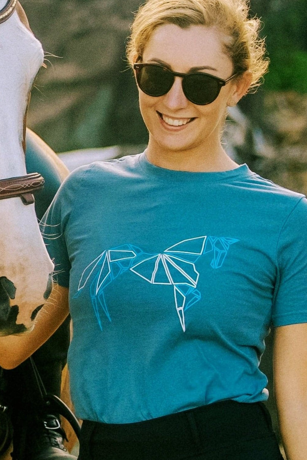 A person with blonde hair wearing sunglasses and a teal Pure Canter Youth PURE Emblem Shirt, featuring a geometric horse design, a true wardrobe staple, stands next to a brown horse with a white stripe on its face. The person is smiling, and the background is outdoors.