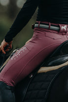 A close-up of a person wearing Pure Canter V1 Fusion Riding Tights - Mulberry with "PURE" and "CANTER" printed on the thigh, riding a black horse. The rider's black attire and the horse's saddle are visible. The rider holds the reins with their left hand, showcasing their equestrian fashion sense.