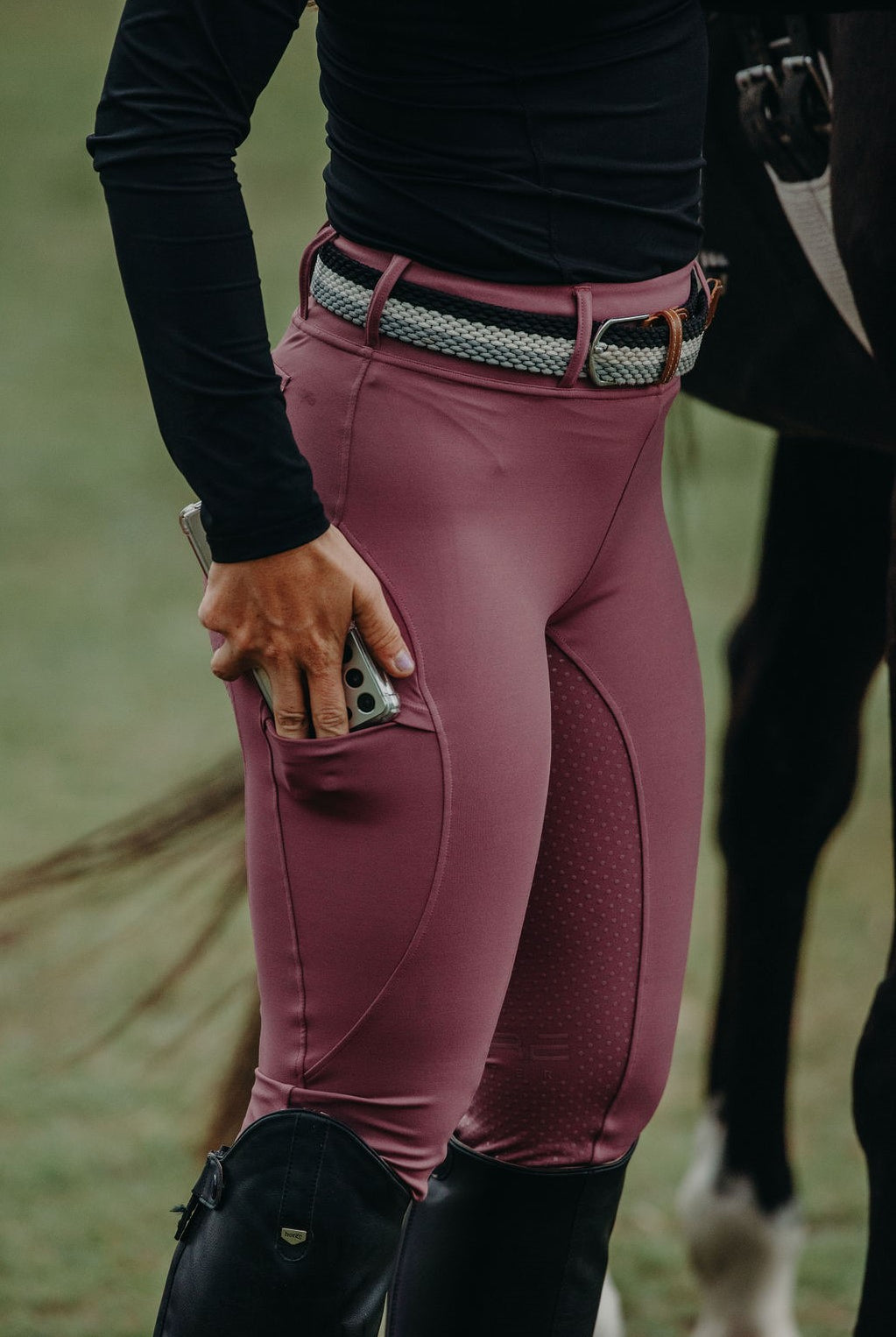 A person wearing Pure Canter Fusion Tights - Mulberry constructed from performance fabric, a woven belt, and black boots holds a partially visible phone in their right hand. The person is standing next to a horse, with the horse's body partially visible in the background.