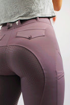 Close-up of a person wearing Pure Canter's V1 Fusion Riding Tights - Plum with buttoned back pockets and a dotted pattern on the seat. The person is in a standing position with their back facing the camera, showcasing the detail and fit of the riding tights made from breathable fabric.