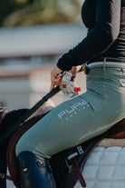 A person wearing a black long-sleeve shirt, light green Fusion Tights - Sage with the text "Pure Canter" on the leg, and black riding boots is sitting on a horse. They are holding reins in their left hand and a whip in their right hand. The horse's saddle and white blanket are visible.