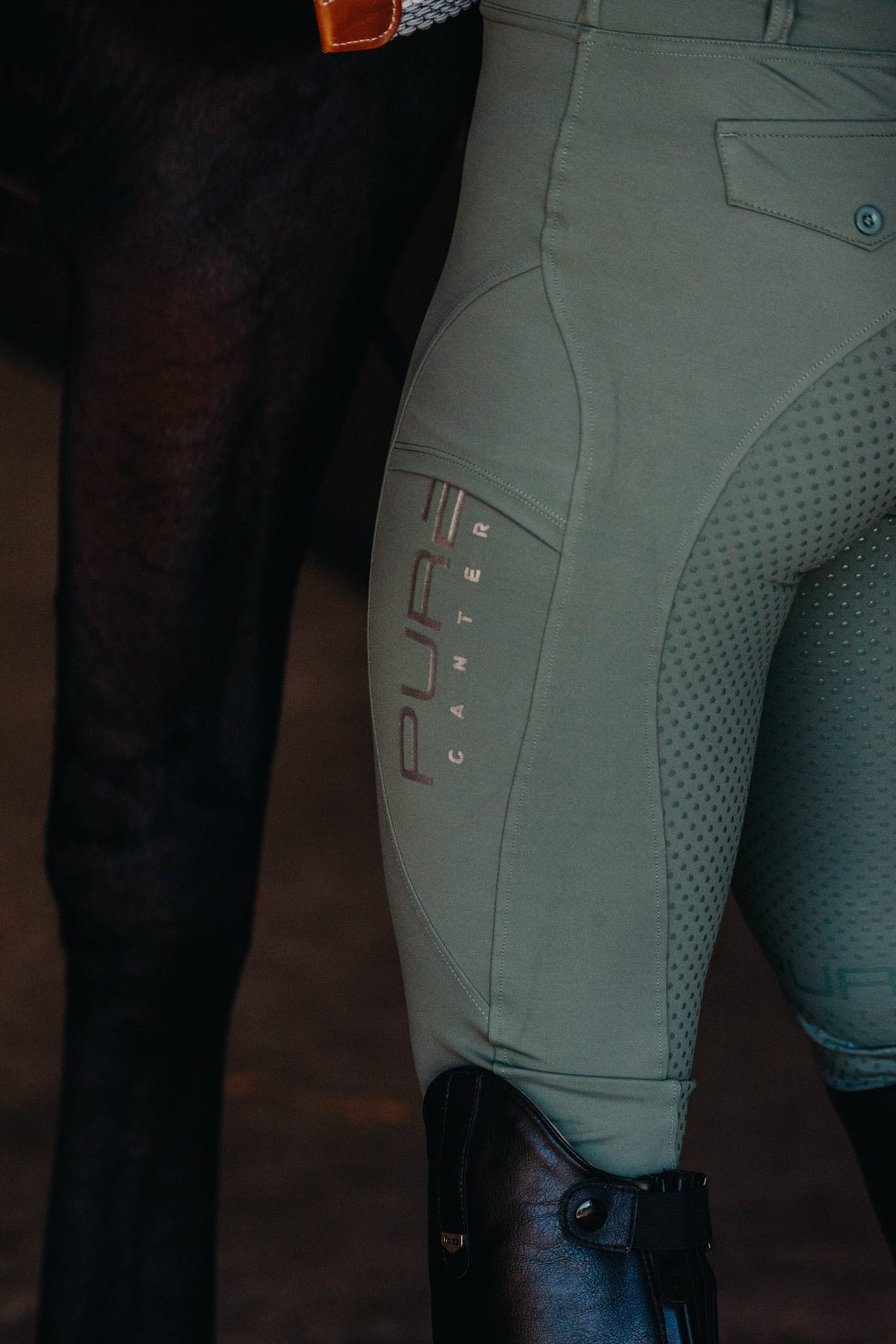 Close-up view of a person wearing beige Fusion Tights - Sage by Pure Canter, showcasing equestrian fashion at its finest. Black riding boots are also partially visible. The dark leg of a horse is seen next to the person.
