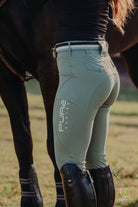 A person dressed in equestrian attire stands beside a horse. They wear light green Fusion Tights - Sage with the brand name "Pure Canter" printed on the side, a black belt, and tall black riding boots. The horse is partially visible, and the setting appears to be outdoors in an elegant display of equestrian fashion.