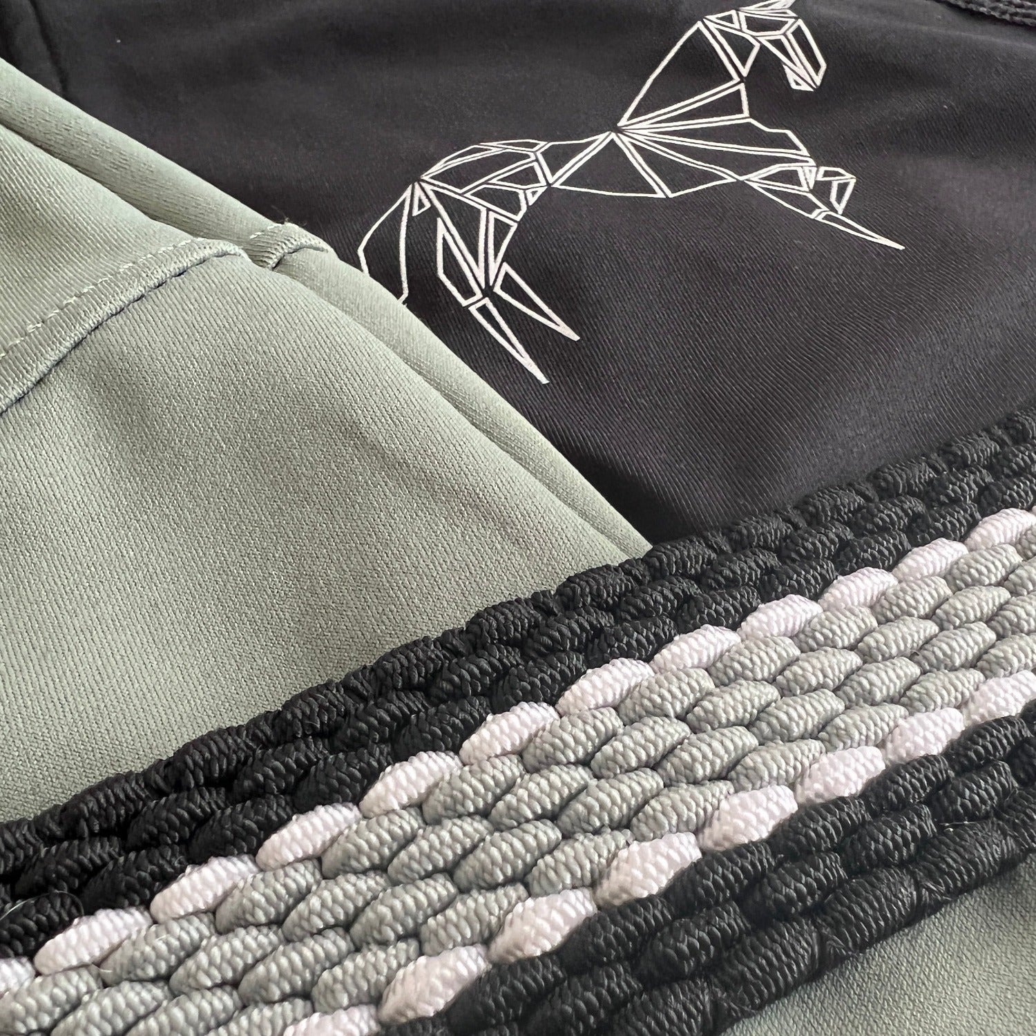 Close-up of black and green fabric with an embroidered geometric horse design on the black section. Below the fabric, Pure Canter's The Braided Belt features a braided strap with interwoven black, gray, and white strands and a secure buckle for added functionality.