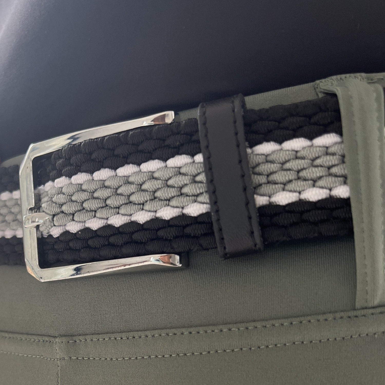 Close-up of Pure Canter's The Braided Belt worn on green pants. The belt features a secure buckle and a pattern consisting of black, gray, and white woven strands with a slight stretch. The metallic silver buckle and black leather loop add a touch of sophistication.