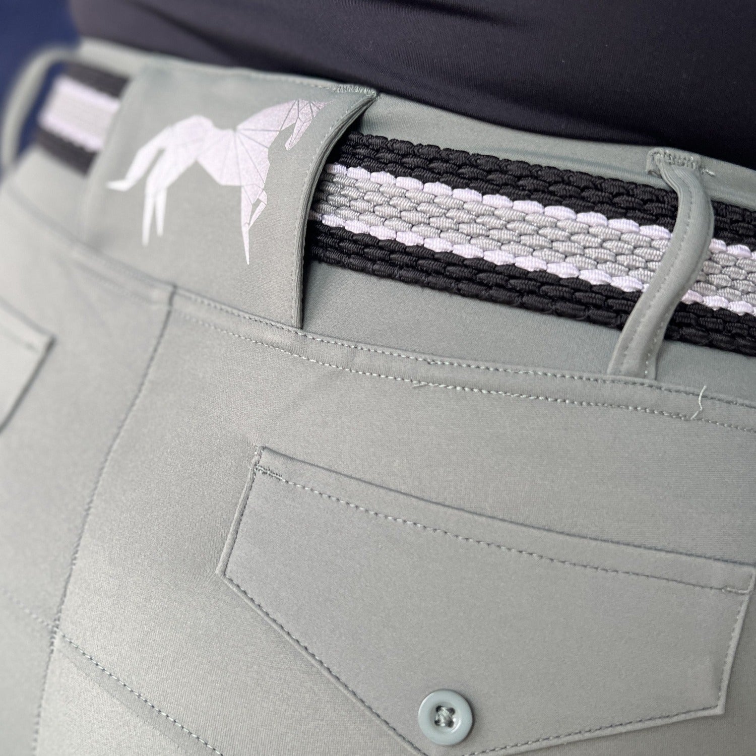 Close-up view of a person wearing light gray pants that have a buttoned back pocket, standing with their back facing the camera. The pants feature The Braided Belt from Pure Canter, showcasing a black and white geometric horse design emblem on the waistband, offering both style and a secure buckle.