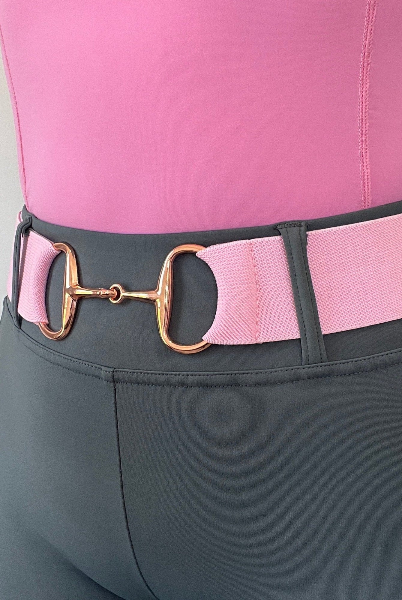 Close-up of a person wearing grey high-waisted pants and a pink shirt. They are accessorized with The Snaffle Belt by Pure Canter, featuring a gold-tone horse bit buckle, ensuring an adjustable fit. The background is plain and light-colored.