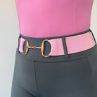 Close-up of a person wearing grey high-waisted pants and a pink shirt. They are accessorized with The Snaffle Belt by Pure Canter, featuring a gold-tone horse bit buckle, ensuring an adjustable fit. The background is plain and light-colored.