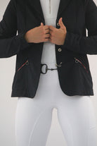 A person in a dark blazer adjusts their jacket. They wear a white shirt underneath and Pure Canter Competition Tights - White with silicone grip patches on the legs and a metallic hook attachment at the waist. The background is a plain, light gray color.