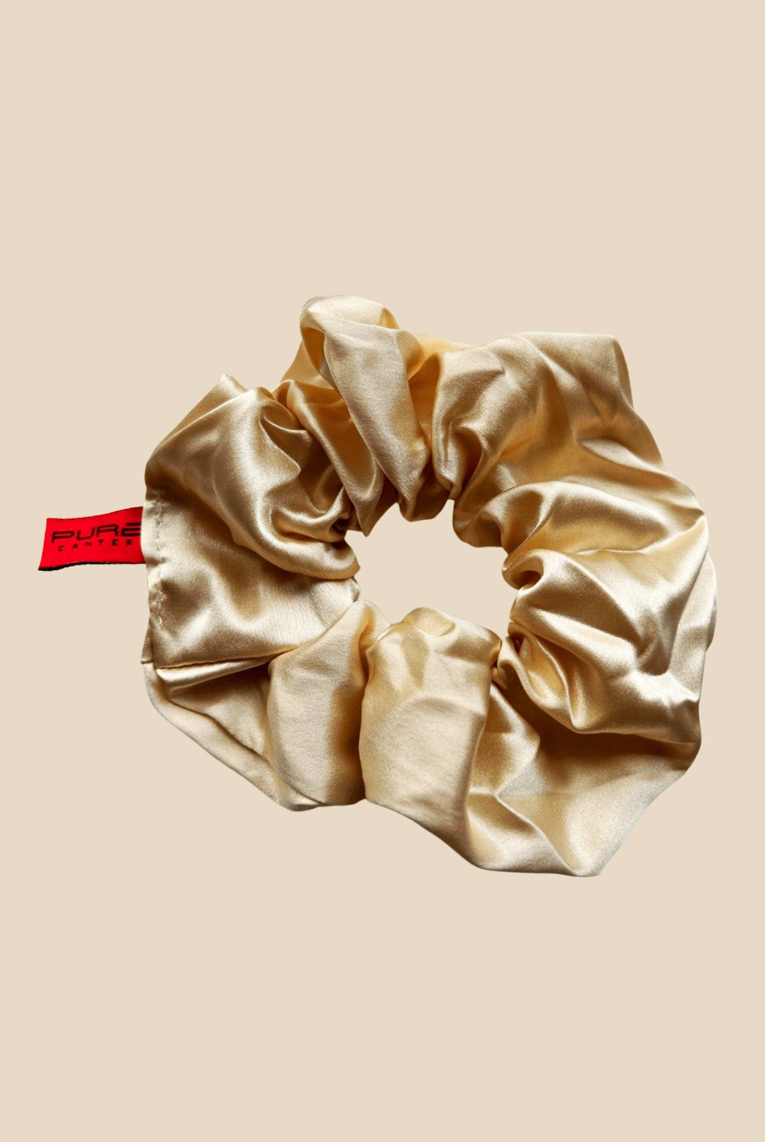 An Oversized Scrunchie by Pure Canter Pty Ltd in beige satin with a slightly glossy finish is displayed against a solid light brown background. It has a red tag with black text attached to one side, making it perfect for casual outings.