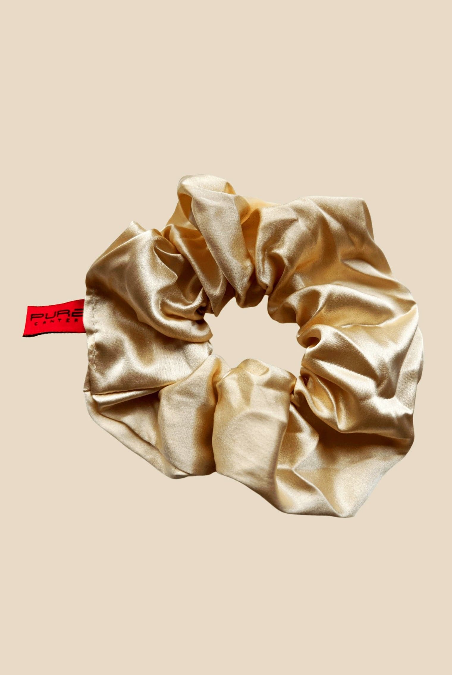 An Oversized Scrunchie by Pure Canter Pty Ltd in beige satin with a slightly glossy finish is displayed against a solid light brown background. It has a red tag with black text attached to one side, making it perfect for casual outings.