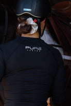 A person wearing a black equestrian helmet and a black long-sleeved shirt from the Pure Canter collection with the text "PURE CANTER" on the back prepares to ride a horse. The person's hair is tied back with a white scrunchie. The background is partially blurred, with hints of a horse and saddle.