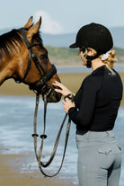 A woman wearing a helmet and a Motion Top Long - Black from the Pure Canter collection stands on a beach, holding a brown horse's reins as she gently strokes its muzzle. The background features sandy shorelines and distant greenery under a clear sky.