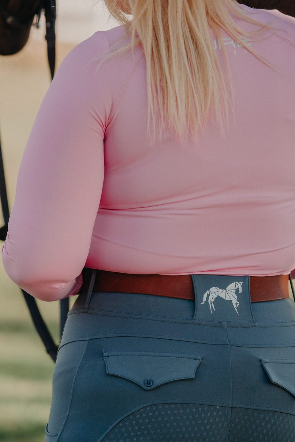 Person wearing a pink, moisture-wicking Pure Canter Motion Top Long - Pink and grey riding pants with a white horse design on the waistband, seen from behind. The person is standing outdoors with light brown hair hanging down.