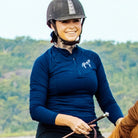 A person wearing a navy blue Pure Canter Motion Top Long, a black helmet, and gloves is smiling while riding a horse. The rider is holding the reins with one hand and a riding crop with the other. A green, wooded background is visible in the distance.