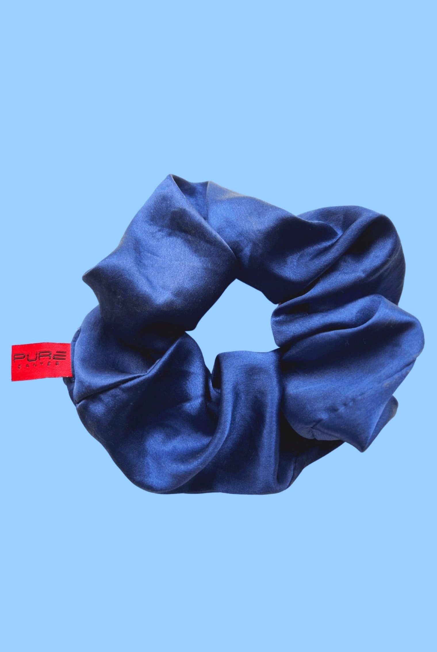 A blue satin Oversized Scrunchie is displayed against a light blue background. The scrunchie, perfect for casual outings, features a red tag with the brand name "Pure Canter Pty Ltd" visible on it.