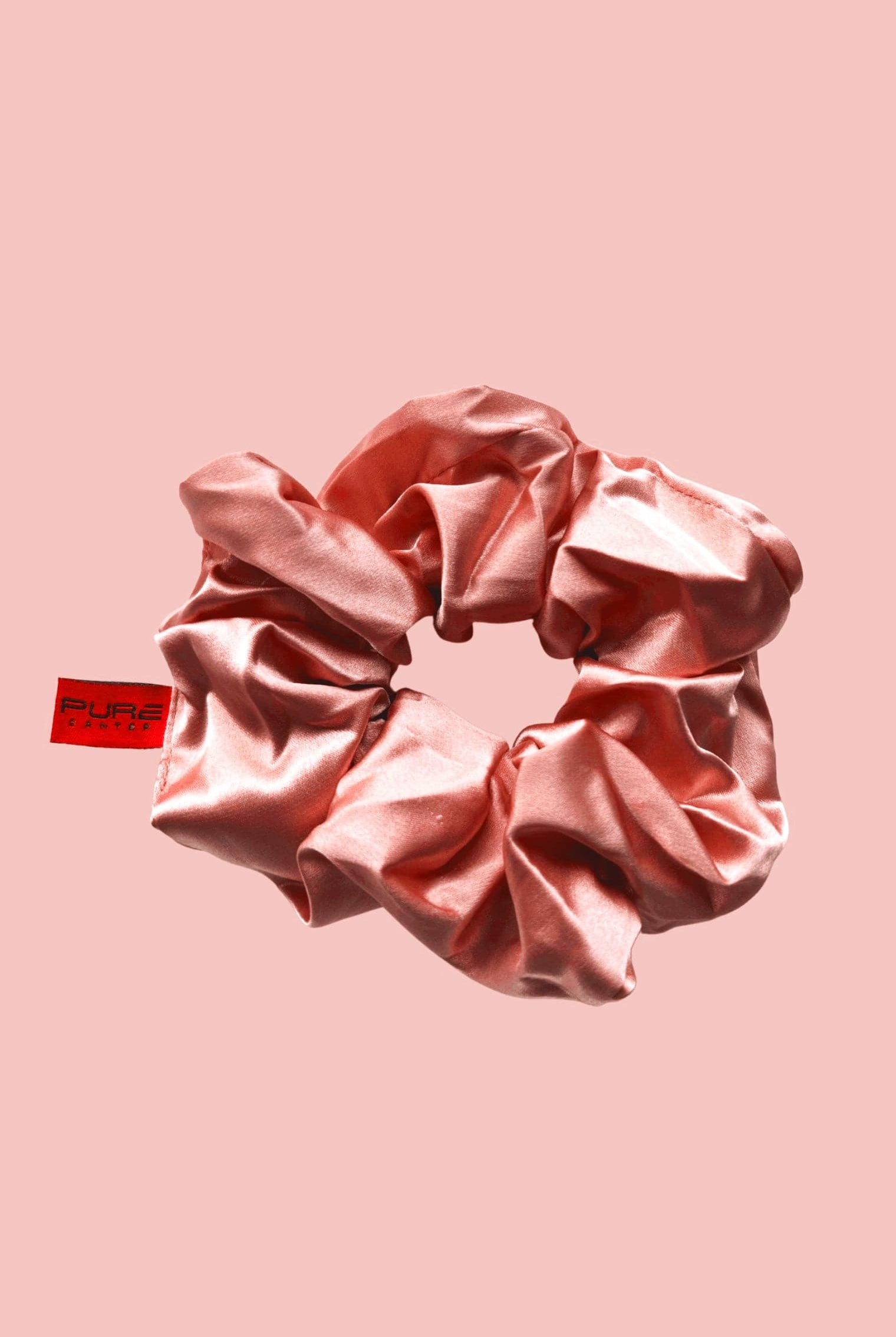 A shiny pink satin Oversized Scrunchies from Pure Canter Pty Ltd with an attached red tag, perfect for casual outings, set against a light pink background.
