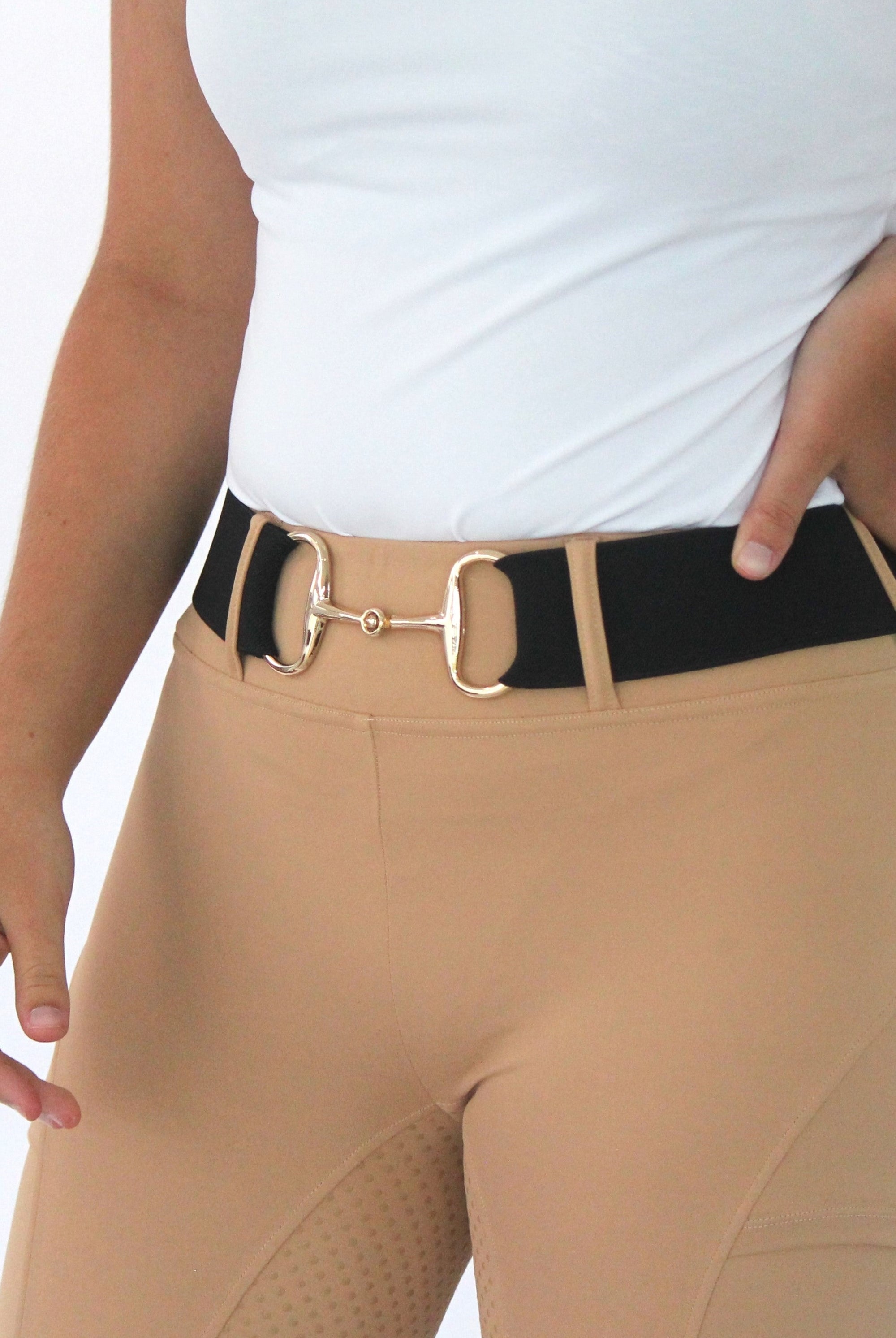 A person wearing tan colored riding pants with a black belt that has a gold horse bit buckle, offering ultimate comfort. Their top is white and tucked into the pants. The person's left hand is resting on their hip, showcasing the adjustable fit of The Snaffle Belt by Pure Canter against a plain white background.