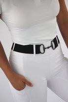 A person is wearing tight white pants and a fitted white shirt, with an elastic stretch belt featuring a large, distinctive buckle at the waist for ultimate comfort. They are wearing The Snaffle Belt by Pure Canter. One hand is casually placed in the pant pocket against a plain white background.