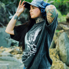 A tattooed person with long dark hair is outdoors, adjusting their hat. They are wearing a super comfy, dark baggy PURE Boyfriend Tee by Pure Canter with a stunning white Pegasus design on it. Rocks and greenery are visible in the background.