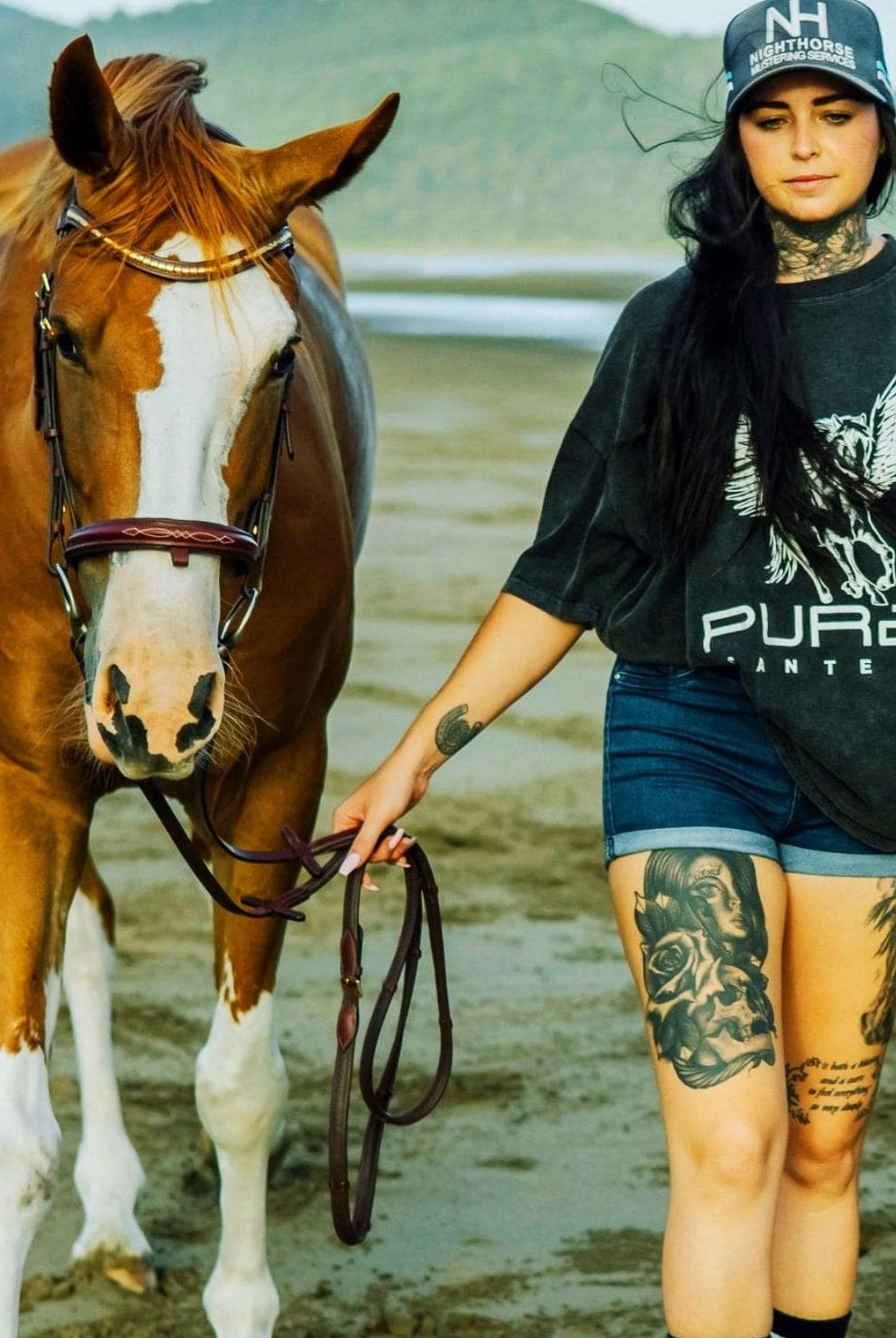 A woman with tattoos holding a brown and white horse's lead rope walks on a beach. She is wearing a black cap, super comfy PURE Boyfriend Tee by Pure Canter with a stunning design, and denim shorts. The background features a grassy landscape and hills under a cloudy sky.