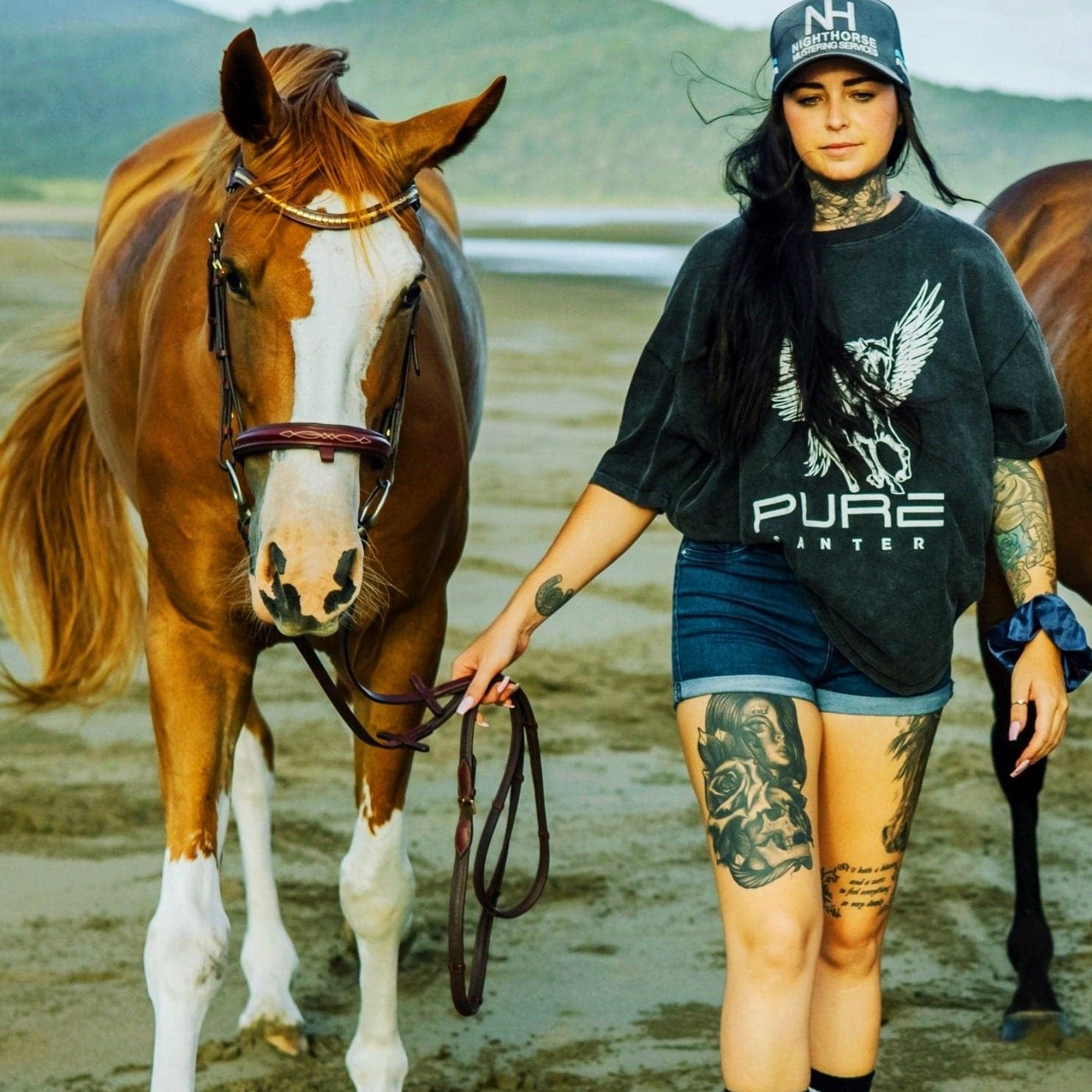 A woman with tattoos holding a brown and white horse's lead rope walks on a beach. She is wearing a black cap, super comfy PURE Boyfriend Tee by Pure Canter with a stunning design, and denim shorts. The background features a grassy landscape and hills under a cloudy sky.