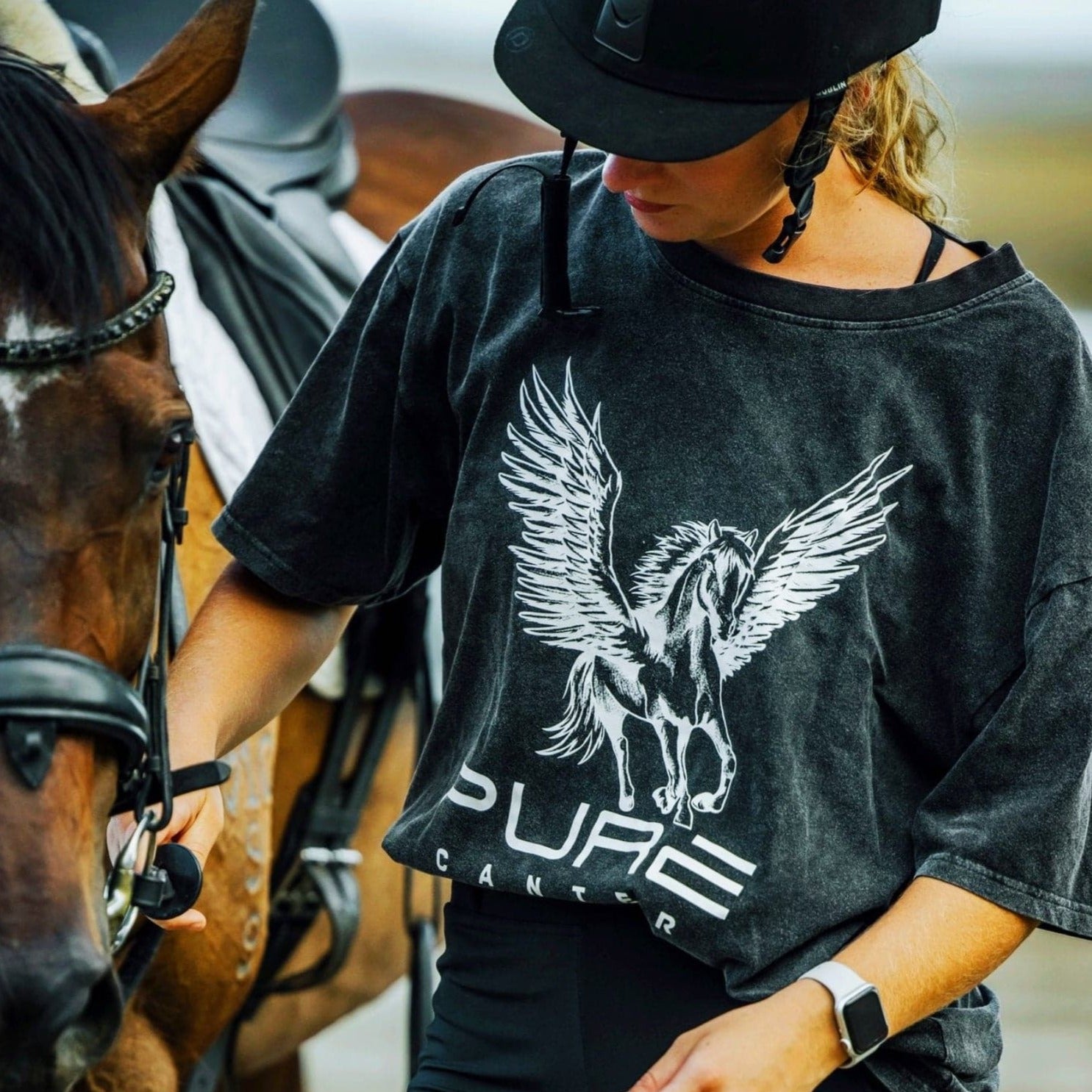 A person wearing a helmet and a baggy Pure Canter PURE Boyfriend Tee with a Pegasus design is leading a horse. The shirt has the text "PURE CANTER." The individual holds the horse's reins and looks downward while standing beside the saddled horse, exuding a relaxed, super comfy vibe.