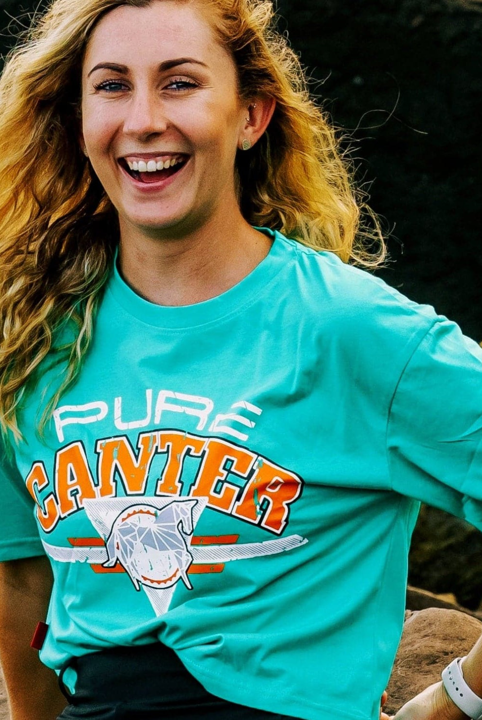 A woman with long, wavy blonde hair is smiling at the camera. She is wearing a teal t-shirt named "Youth PURE Sports Crop" from Pure Canter Pty Ltd, with the words "PURE CANTER" and a graphic of a horse, paired perfectly with high-waisted pants. She is outdoors with large rocks in the background.