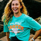 A woman with long, wavy blonde hair is smiling at the camera. She is wearing a teal t-shirt named "Youth PURE Sports Crop" from Pure Canter Pty Ltd, with the words "PURE CANTER" and a graphic of a horse, paired perfectly with high-waisted pants. She is outdoors with large rocks in the background.