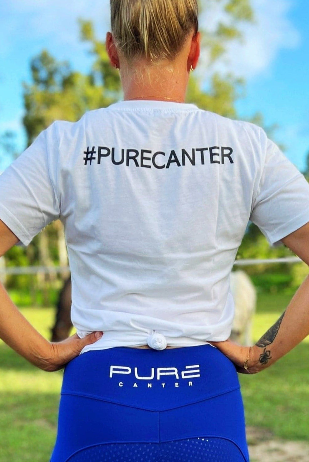 A person stands outdoors with their back to the camera, hands on hips. They sport a white Youth PURE Emblem Shirt by Pure Canter with the hashtag "#PURECANTER" and blue leggings featuring "PURE" on the waistband—true wardrobe staples. Lush green trees and a clear blue sky form the backdrop.