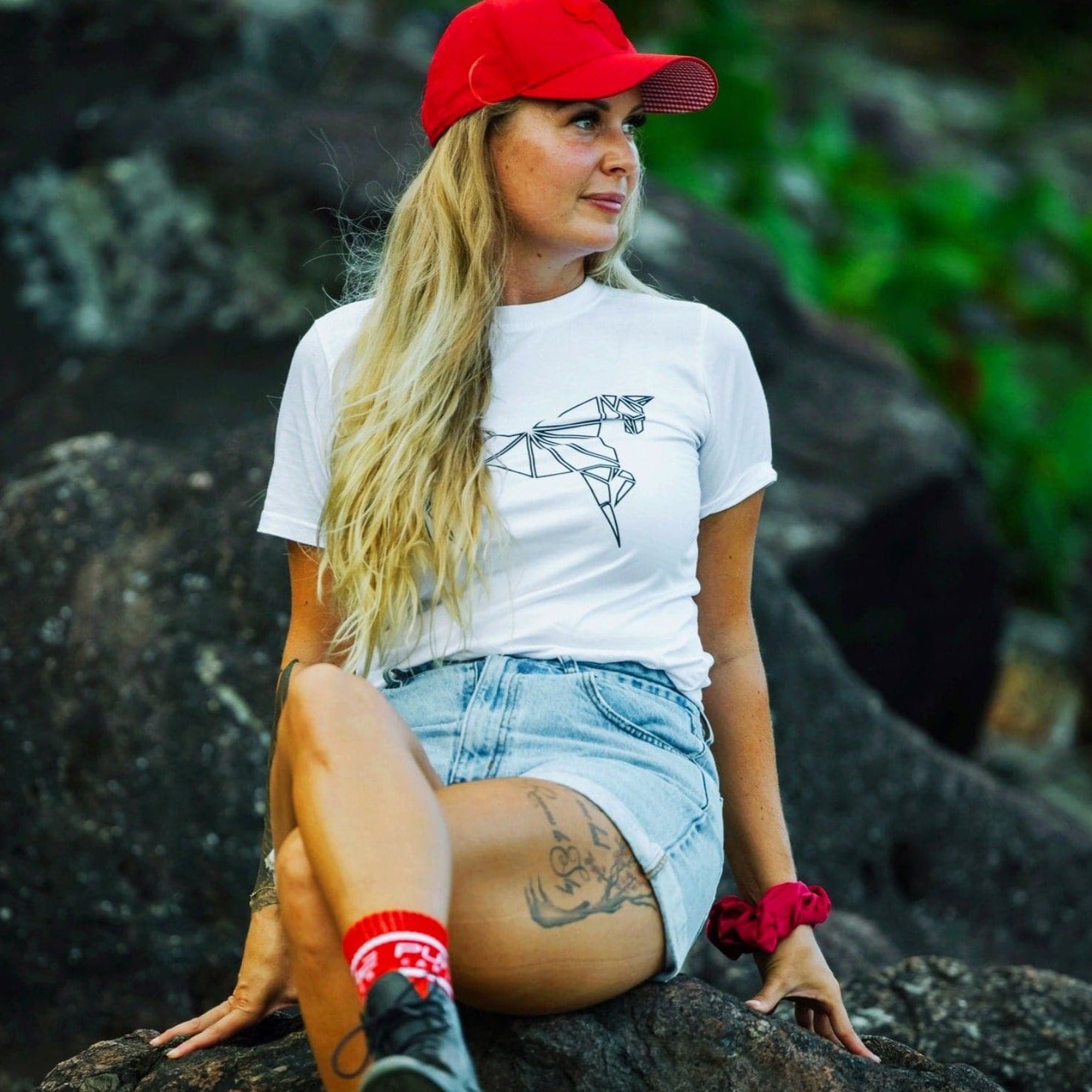 A woman with long blonde hair wearing a red cap sits on a rock. She is dressed in a white Pure Canter Youth PURE Emblem Shirt, blue denim shorts, and red socks. She has a tattoo on her left thigh and a red scrunchie on her left wrist. The background is blurred greenery, creating an idyllic scene.