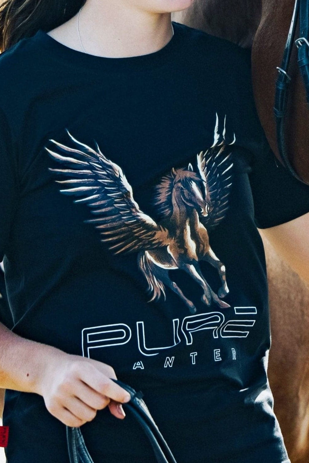 A person wearing a black t-shirt with a detailed graphic of a winged horse and the text "PURE" and "ANTER." The Youth PURE Wings Tee, a product from Pure Canter Pty Ltd, featuring a statement design, runs very small. The individual holds the reins of a brown horse standing beside them. Both are partially visible, suggesting an outdoor environment.