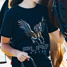 A person wearing a black t-shirt with a detailed graphic of a winged horse and the text "PURE" and "ANTER." The Youth PURE Wings Tee, a product from Pure Canter Pty Ltd, featuring a statement design, runs very small. The individual holds the reins of a brown horse standing beside them. Both are partially visible, suggesting an outdoor environment.