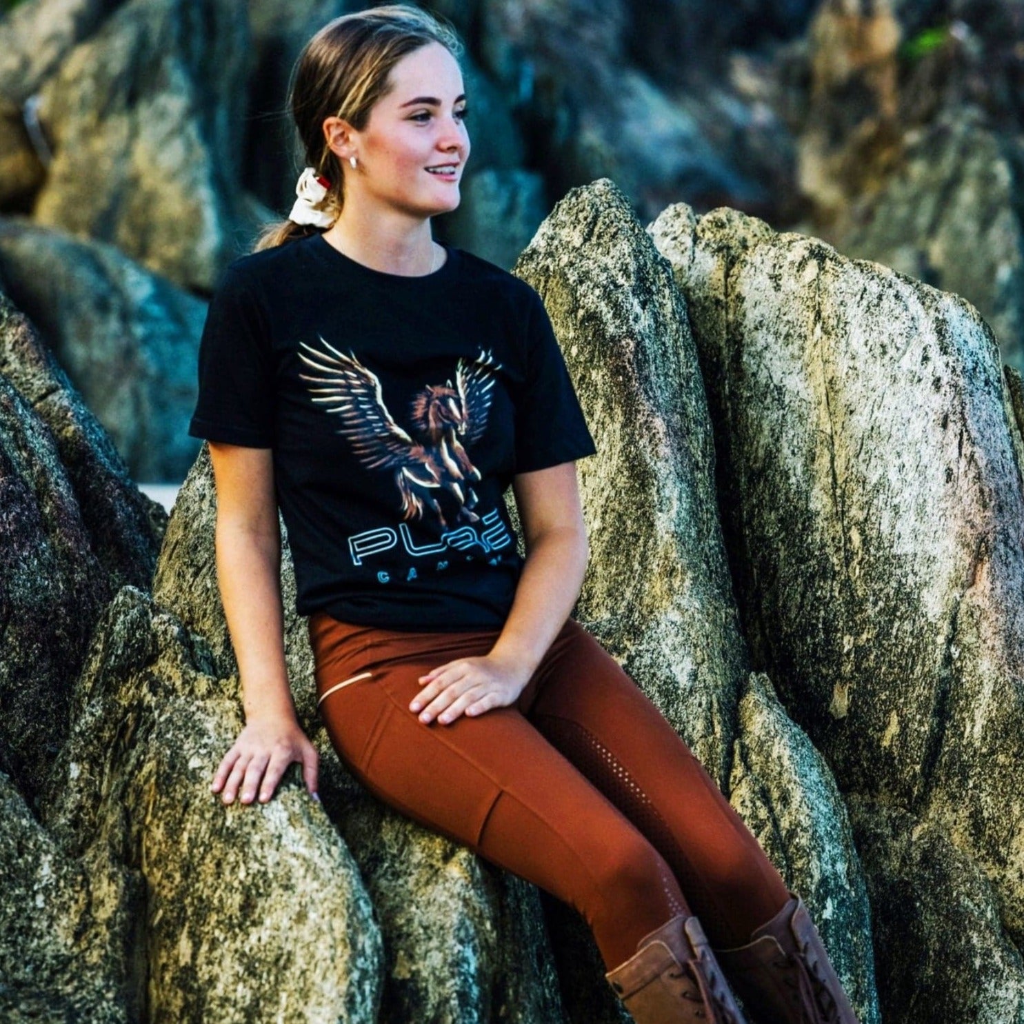 A person with long hair sits on large rocks outdoors, wearing a black Youth PURE Wings Tee from Pure Canter Pty Ltd with a graphic of a winged horse and the word "PLACE." They have brown pants and tall brown boots. The background features more large, rough-textured rocks. The person is looking to the side, smiling.